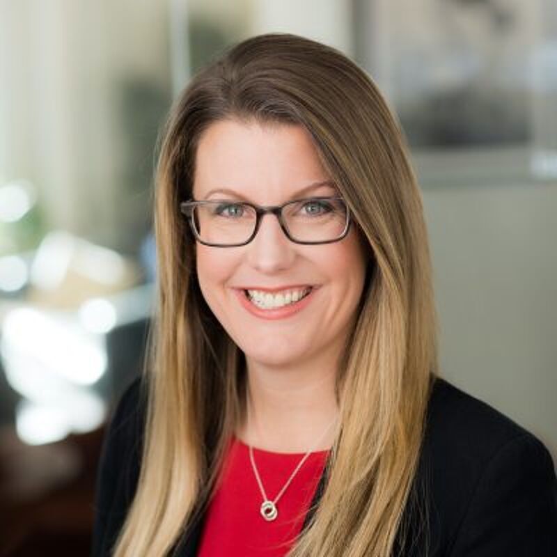 The image shows a woman with long blonde hair, wearing glasses, a red top, and a black blazer. She is smiling at the camera. She has a necklace with a pendant. The background is blurred, suggesting a professional setting.